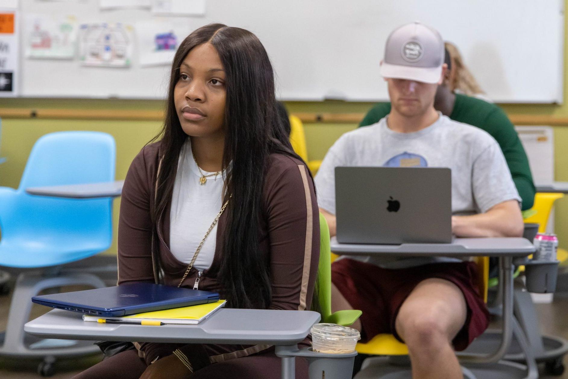 A high school girl sitting at a desk in a classroom with other students sitting at desks behind her.