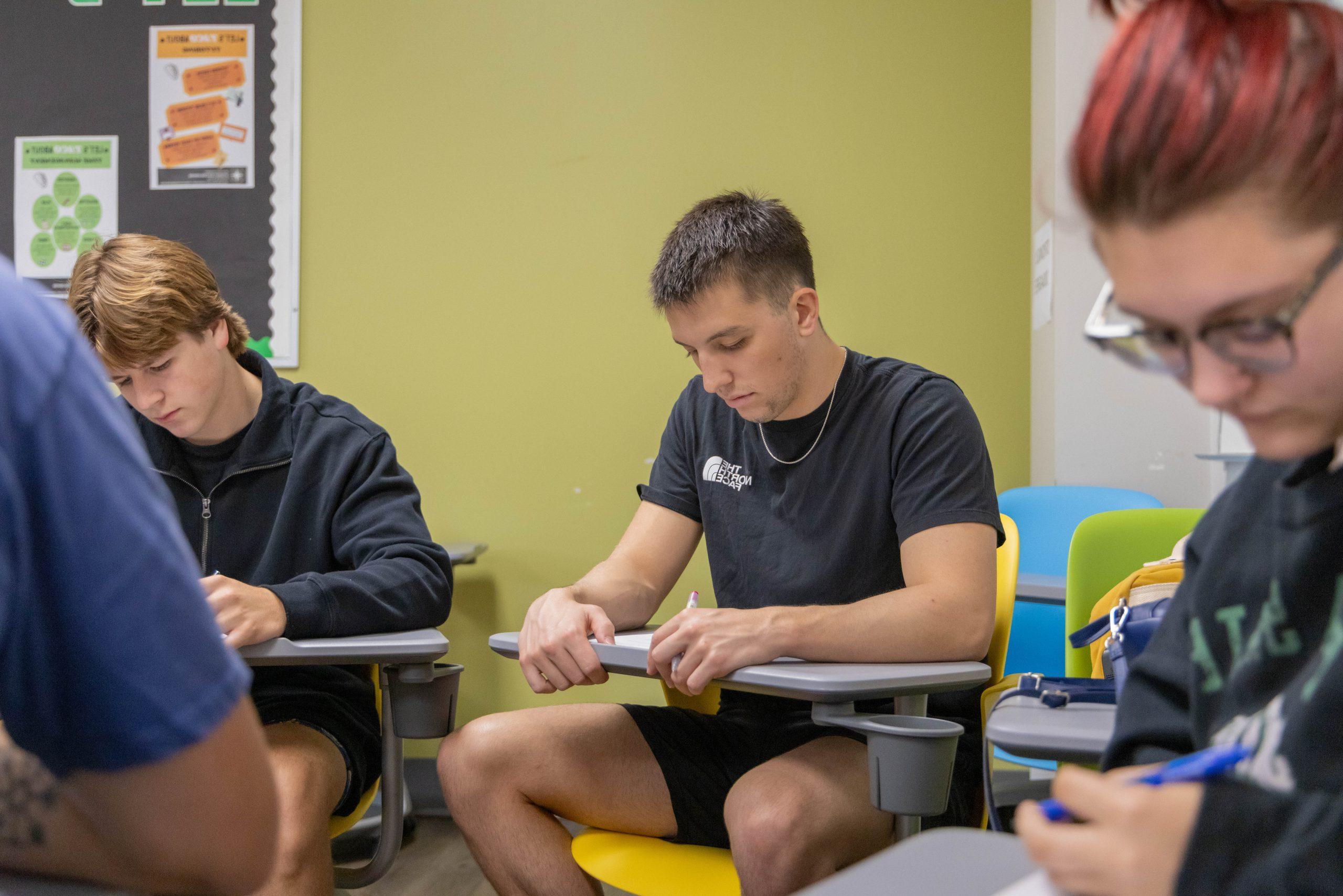 Three high school students sitting at desks in a classroom looking down at papers.