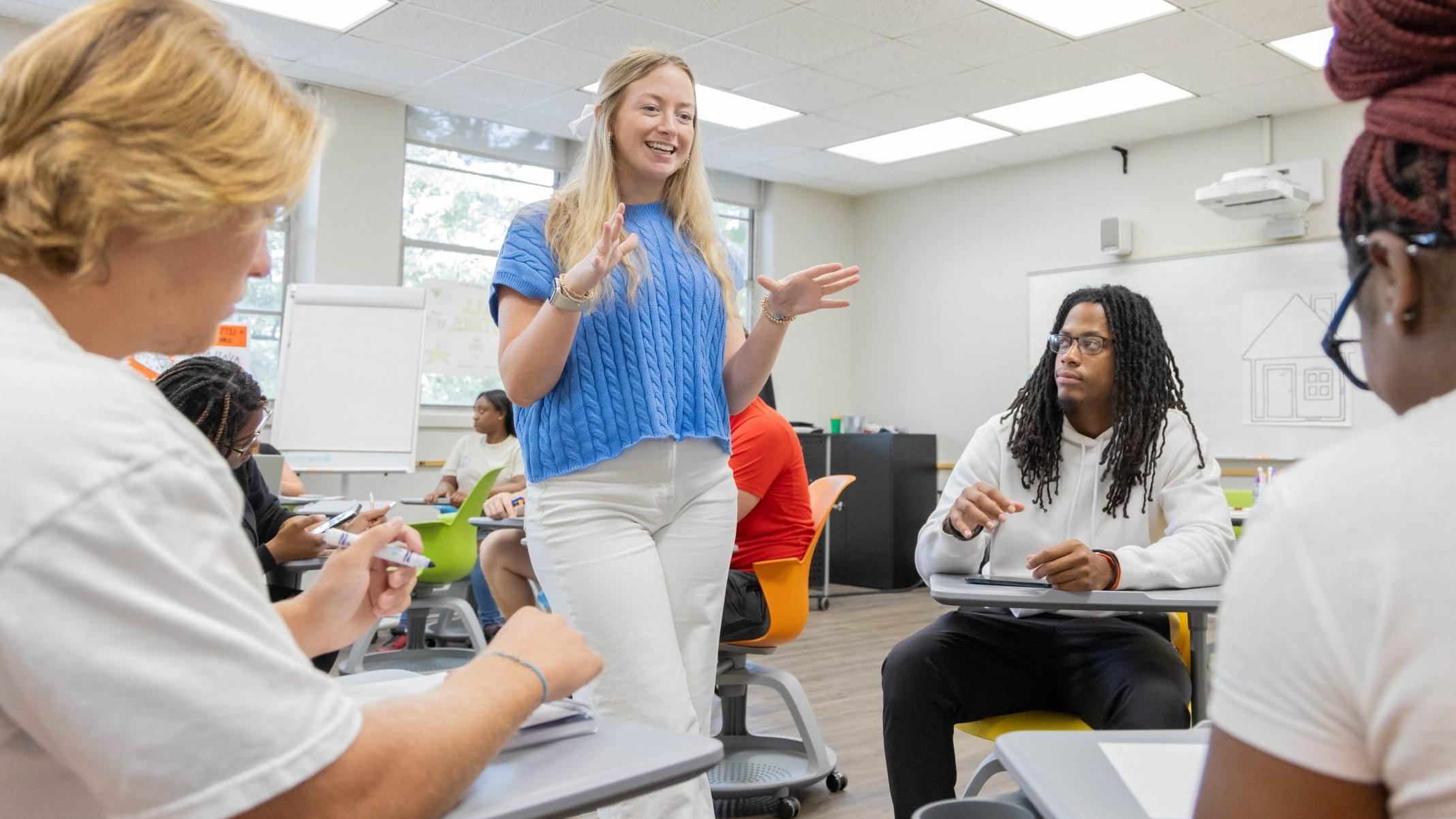 A teacher standing in front of a small group of three students who are sitting at desks in a classroom.